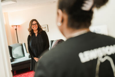 A woman in glasses stands and smiles in a living room, facing another person whose back is to the camera, suggesting a warm and welcoming atmosphere like that found at a counseling center. Saint Joseph's College of Maine