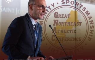 A man in a suit speaks at a podium with the "Great Northeast Athletic Conference" logo in the background. Text overlay reads "Executive of the Year." Brian Curtin receives his award during the GNAC Awards ceremony. Saint Joseph's College of Maine