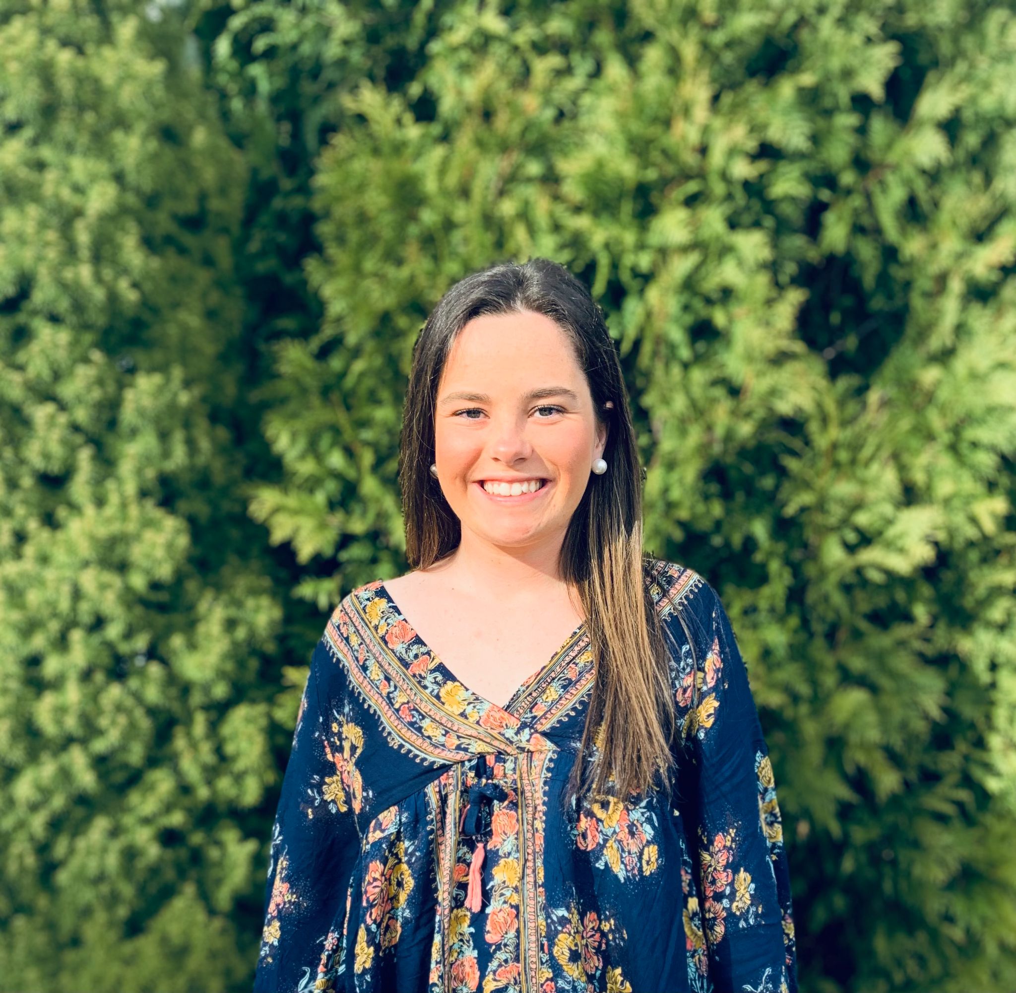A woman stands outside in front of green foliage, smiling. She is wearing a dark floral-patterned top, looking ready for another empowering session at the Career Studio. Saint Joseph's College of Maine