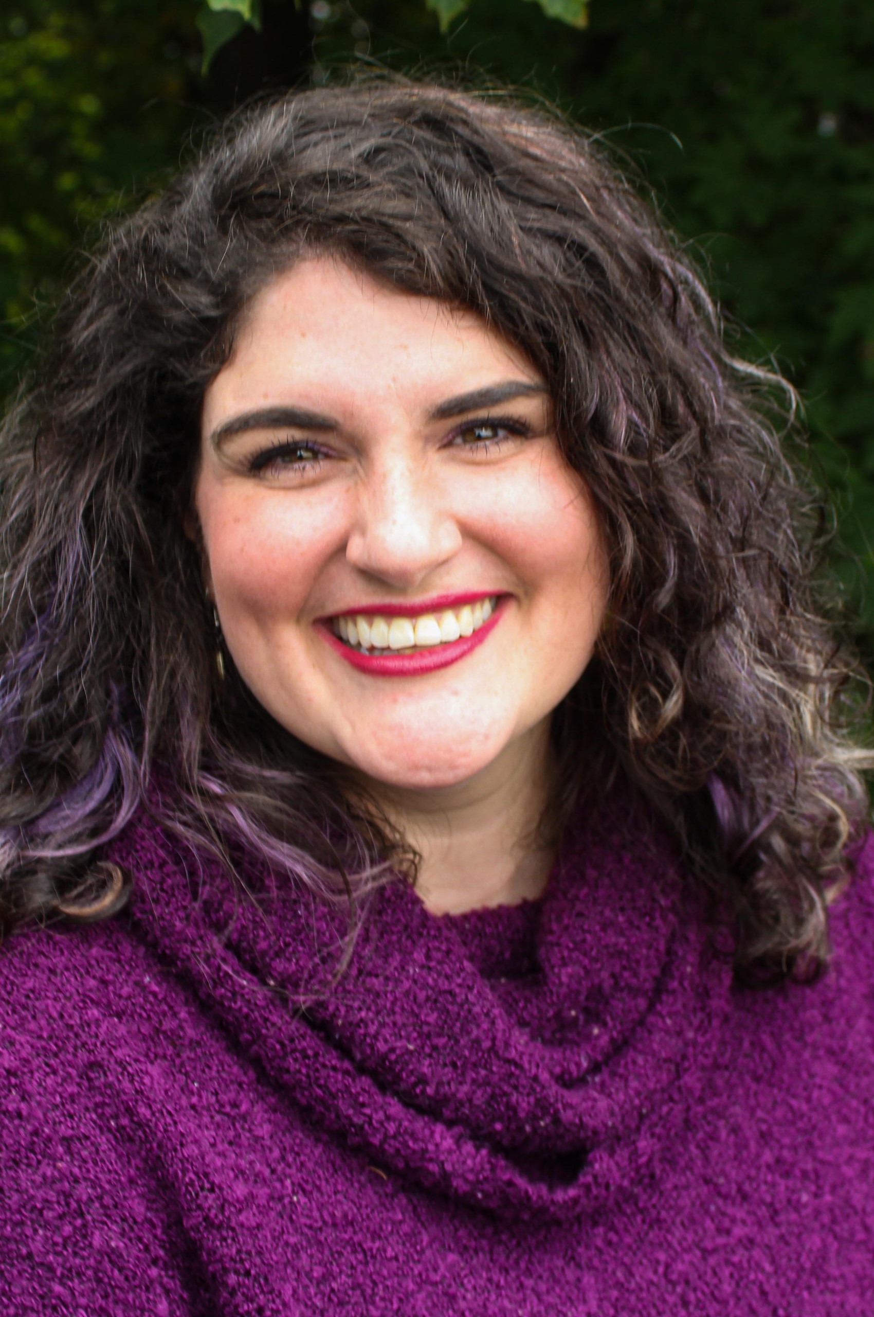Carly Brownsberger Headshot 2 Woman with curly dark hair wearing a purple sweater smiles at the camera, standing outdoors with greenery in the background, reflecting confidence from her recent Career Studio career development experience. Saint Joseph's College of Maine