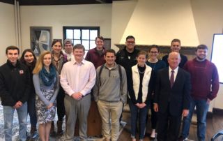 A group of people, including ten young adults and one older man in a suit, stand indoors on campus in front of a fireplace, smiling at the camera. The older man, former U.S. Assistant Secretary of State Philip J. Crowley, stands proudly among the students. Saint Joseph's College of Maine