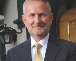 A man with short light hair and a beard, wearing a dark suit, white shirt, and yellow tie, stands outdoors smiling. Behind him is a building featuring a lantern. James Dlugos of the D3 Presidents Advisory Group appears confident in his presidential role. Saint Joseph's College of Maine