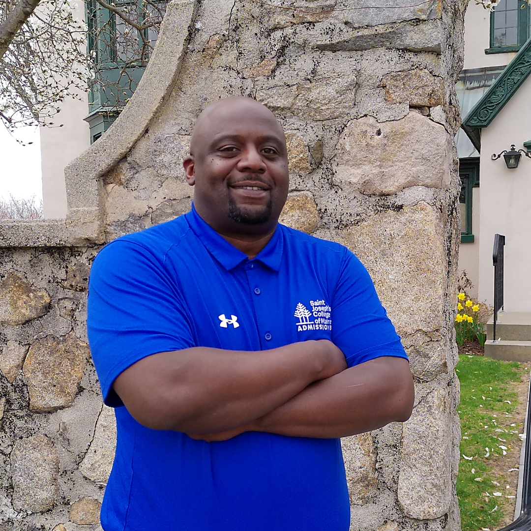 Kareem Photo A man in a blue Saint Joseph's College Admissions On Campus polo shirt stands with arms crossed in front of a stone wall and building. Saint Joseph's College of Maine