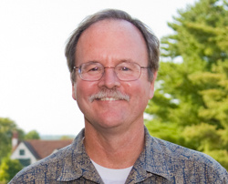 A man with glasses and a mustache, Professor Michael Connolly, smiles at the camera, standing outdoors with trees and houses in the background. Saint Joseph's College of Maine