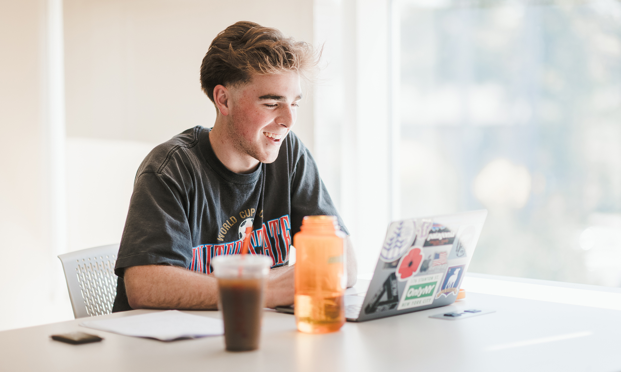 A young man sits at a desk, smiling while using a laptop covered in stickers. A large water bottle and a cup of iced coffee are on the desk. Sunlight streams through the window in the background. Saint Joseph's College of Maine