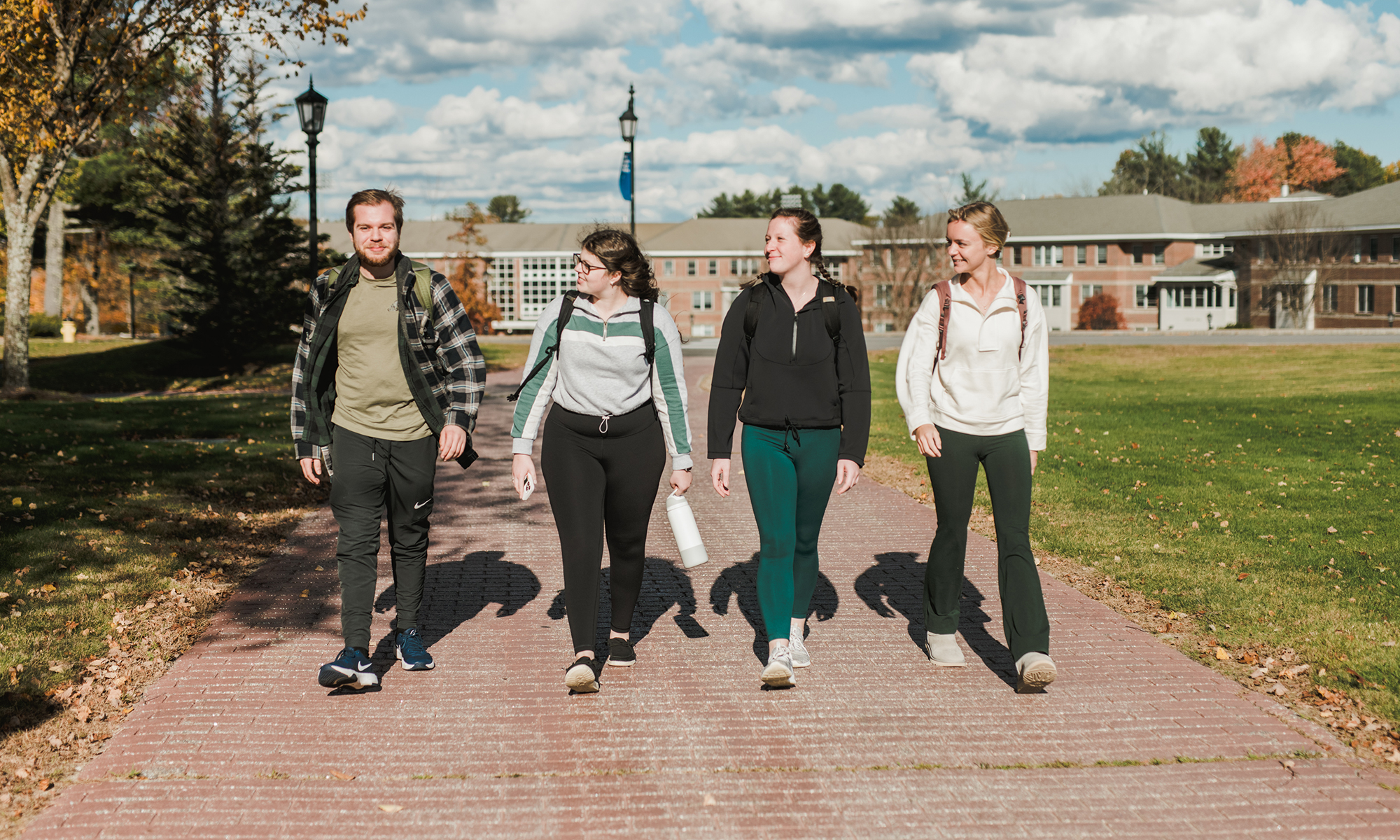 Four people walk side by side on a brick pathway through a green campus area with trees and a building in the background. Saint Joseph's College of Maine