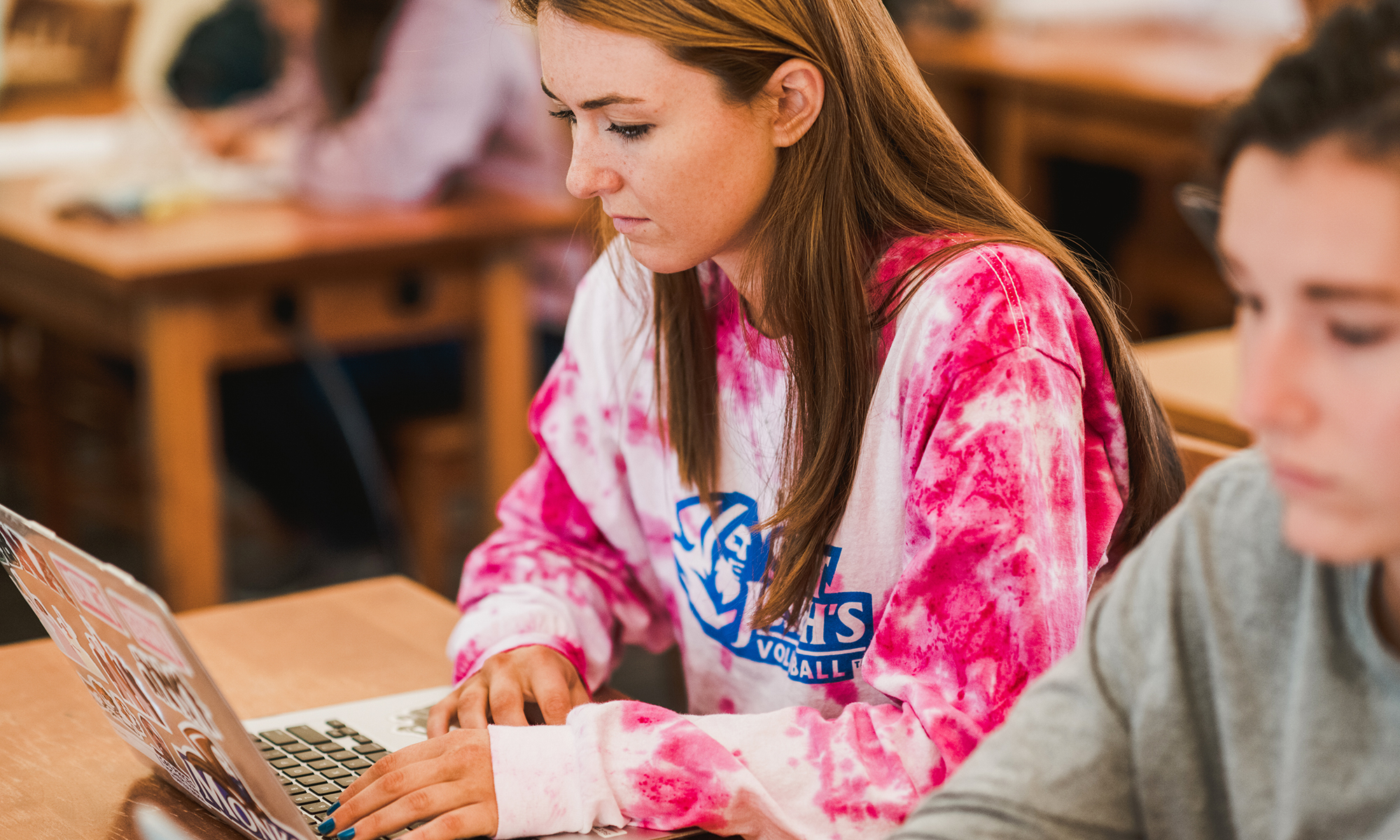 A person in a pink tie-dye shirt uses a laptop at a desk in a classroom setting. Saint Joseph's College of Maine