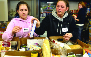 Two women, wearing name tags reading "Rachel" and "Julie", sort food items in boxes at a food bank. Reflections of their commitment glimmer as they work diligently. Another person in the background also handles food items, ensuring no cupboard is ever empty. Saint Joseph's College of Maine