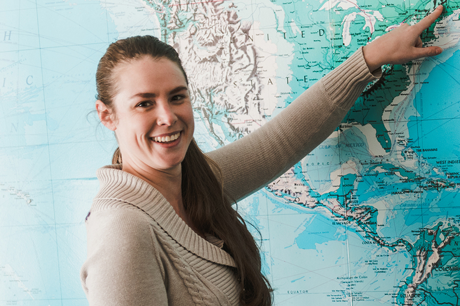 A woman with a ponytail, smiling and pointing to a location in the northeastern United States, stands in front of a large map. She is eager to share that this area hosts an esteemed graduate program available both on-campus and as an online program. Saint Joseph's College of Maine