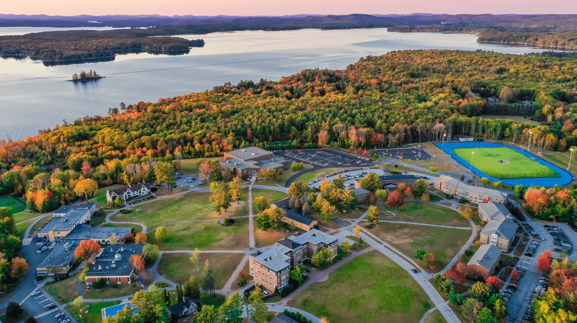 Aerial view of a university campus in the fall, surrounded by colorful trees and situated near a large lake. Buildings, pathways, a sports field, and parking lots are visible. Saint Joseph's College of Maine