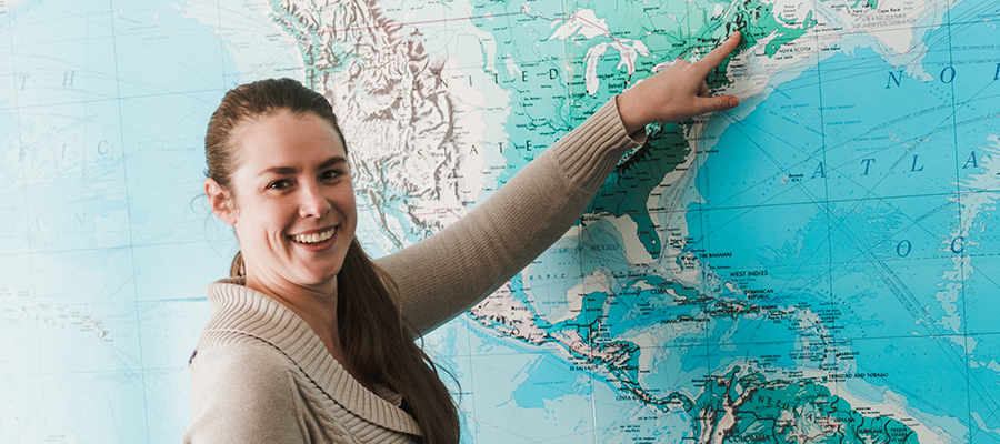 A woman, smiling in a beige sweater, points at a location on a large world map. The background showcases SJC Online's redesign. Saint Joseph's College of Maine