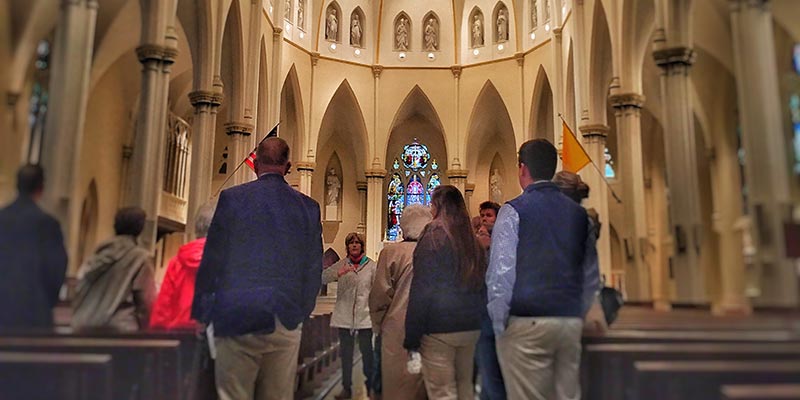 A group of people stands inside a large church with high arches and stained glass windows, facing a guide from the Center for Faith and Spirituality who is speaking in front of them. Saint Joseph's College of Maine