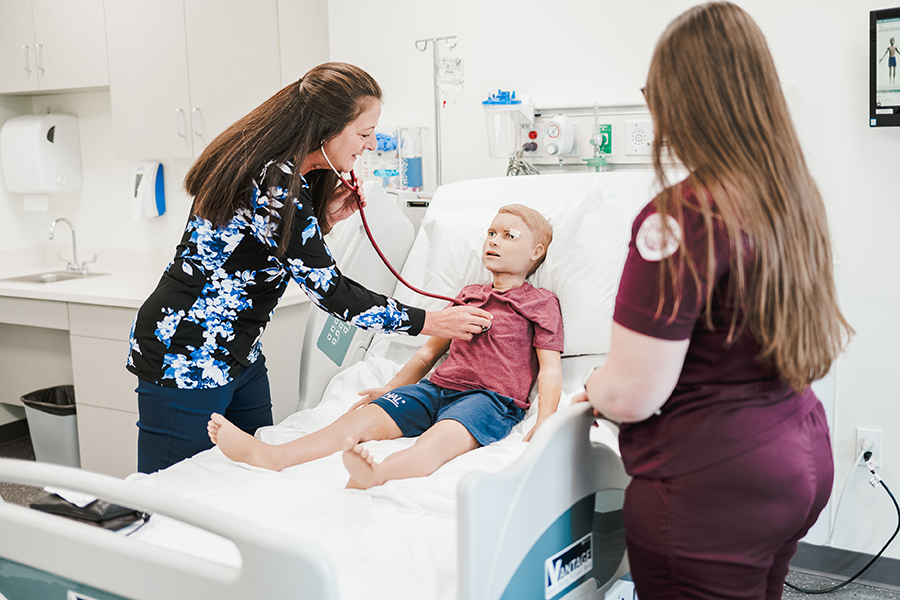 A healthcare provider, demonstrating the value of partnerships in patient care, uses a stethoscope to listen to the chest of a young patient sitting on a hospital bed while another healthcare worker stands nearby. Saint Joseph's College of Maine