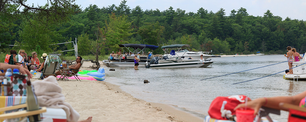People relax on a sandy beach with trees in the background at the Sebago Lake Symposium. Several boats are anchored near the shore, and some individuals are wading in the water. Beach chairs and floaties are scattered around. Saint Joseph's College of Maine